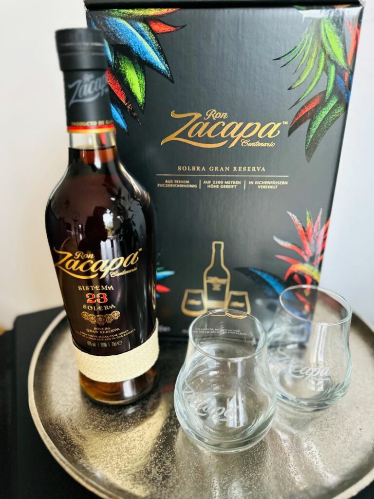 Eine Flasche Ron Zacapa Centenario 23 Jahre mit zwei Gläsern von Ron Zacapa und der Geschenkverpackung im Hintergrund.